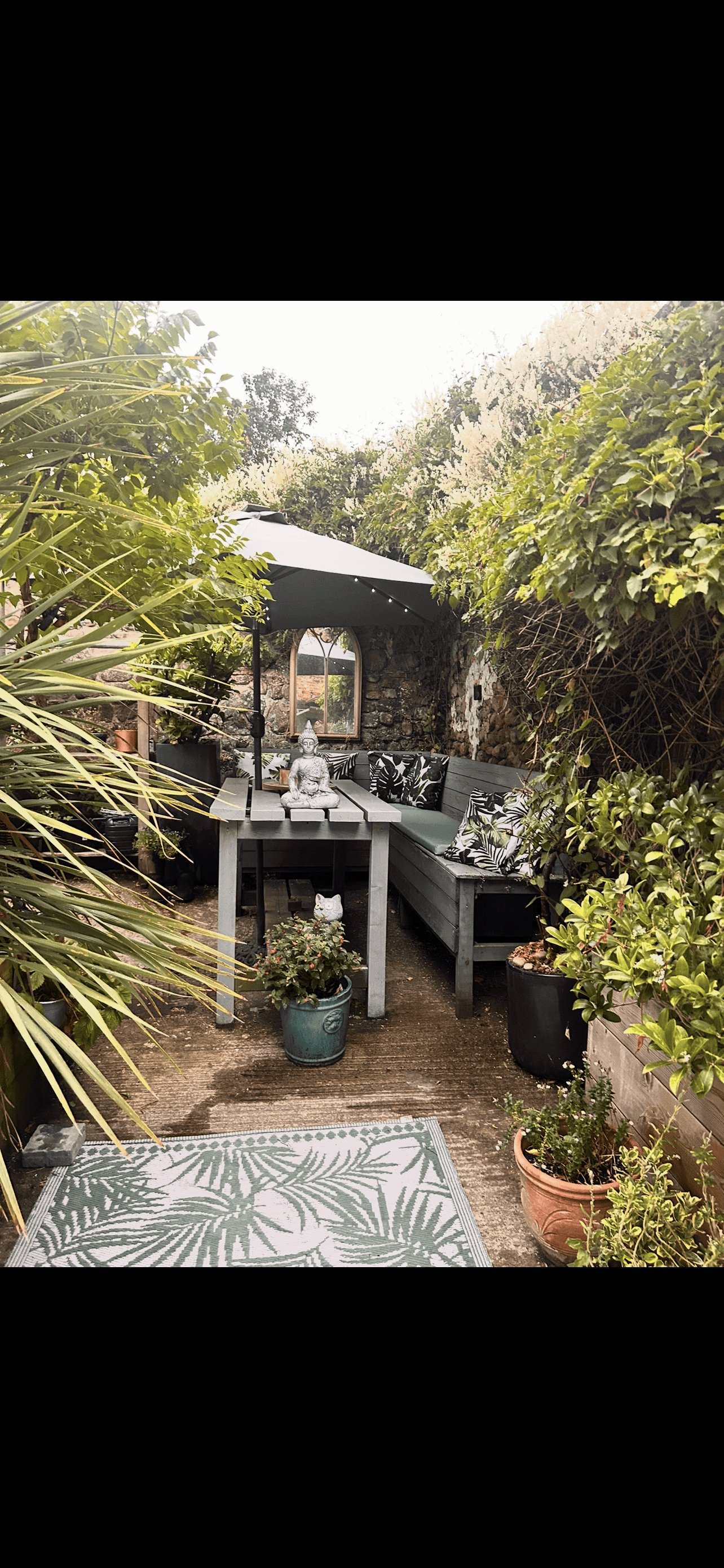 Cozy garden patio with grey wooden furniture, tropical cushions, and lush greenery under an umbrella.