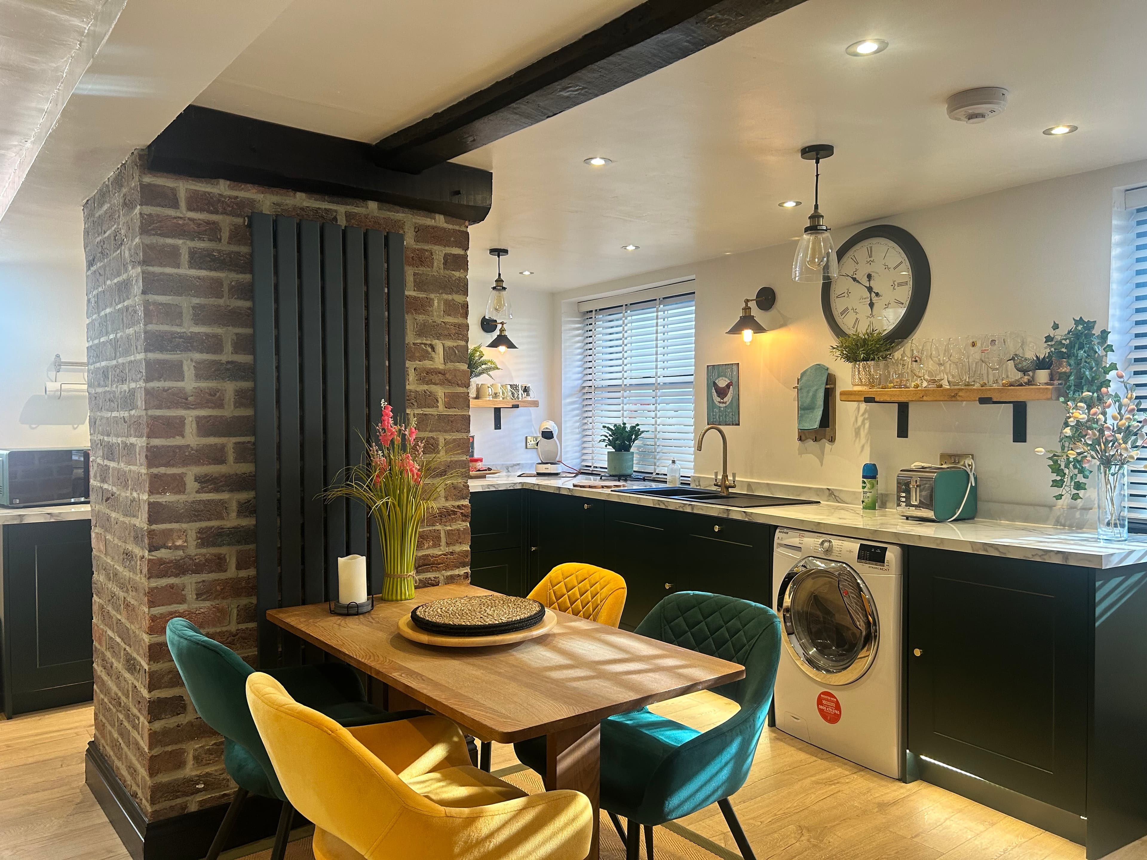 Modern kitchen featuring dark green cabinets and a wooden dining table with colorful velvet chairs.