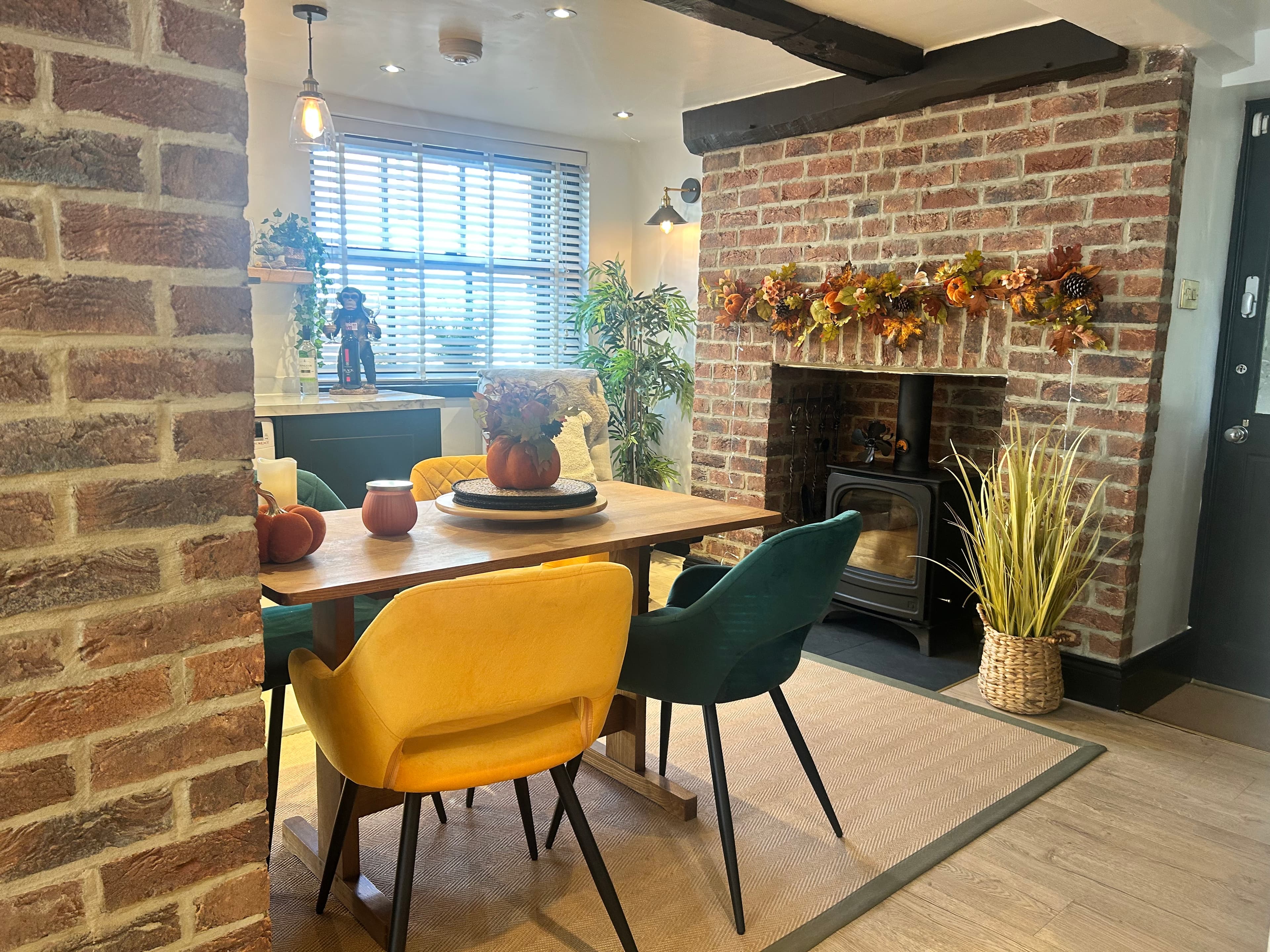 Wooden dining table with yellow and teal chairs beside a brick fireplace with autumn decor.