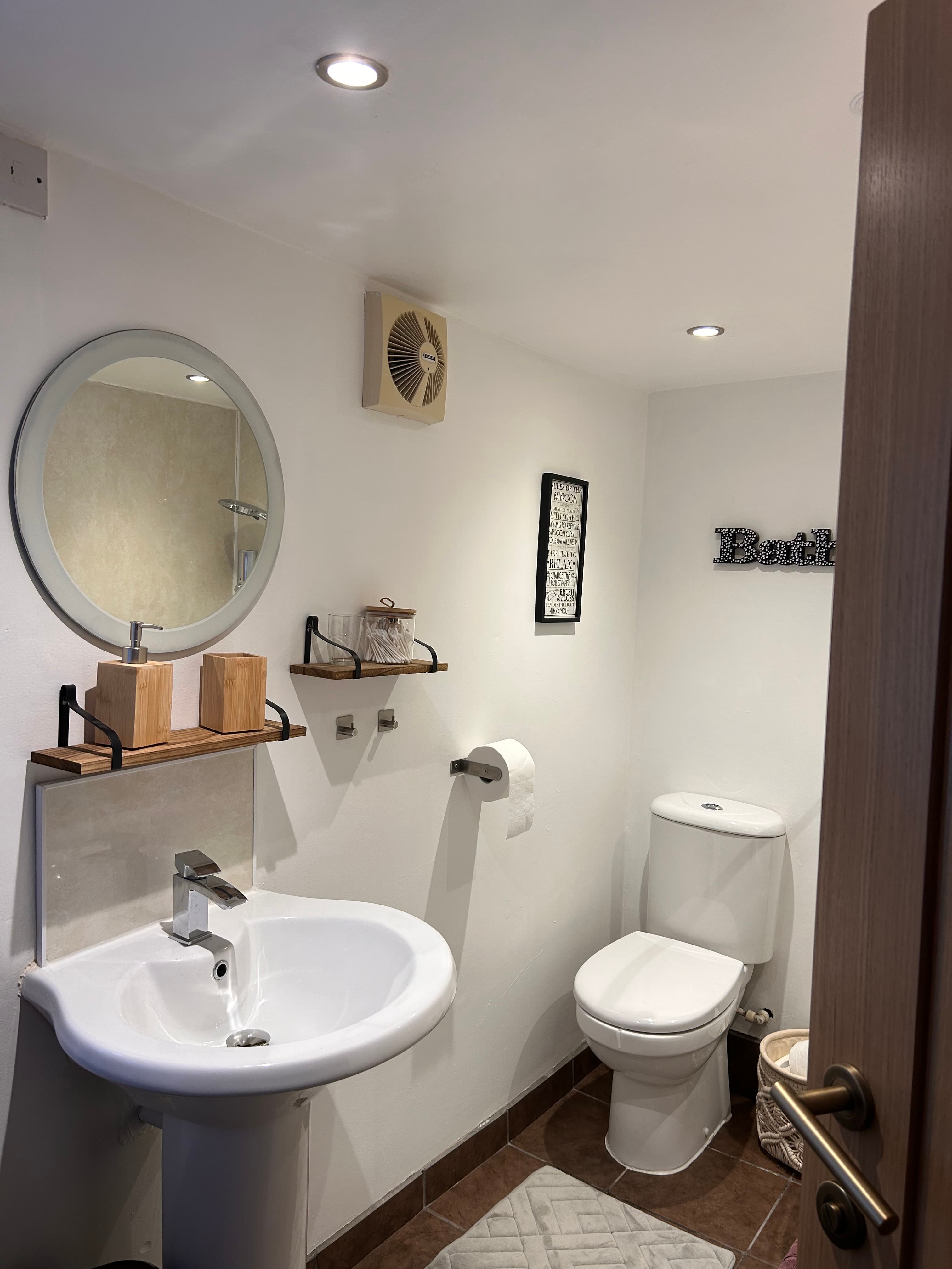 Small white bathroom featuring a pedestal sink, round mirror, wooden shelves, and a toilet.