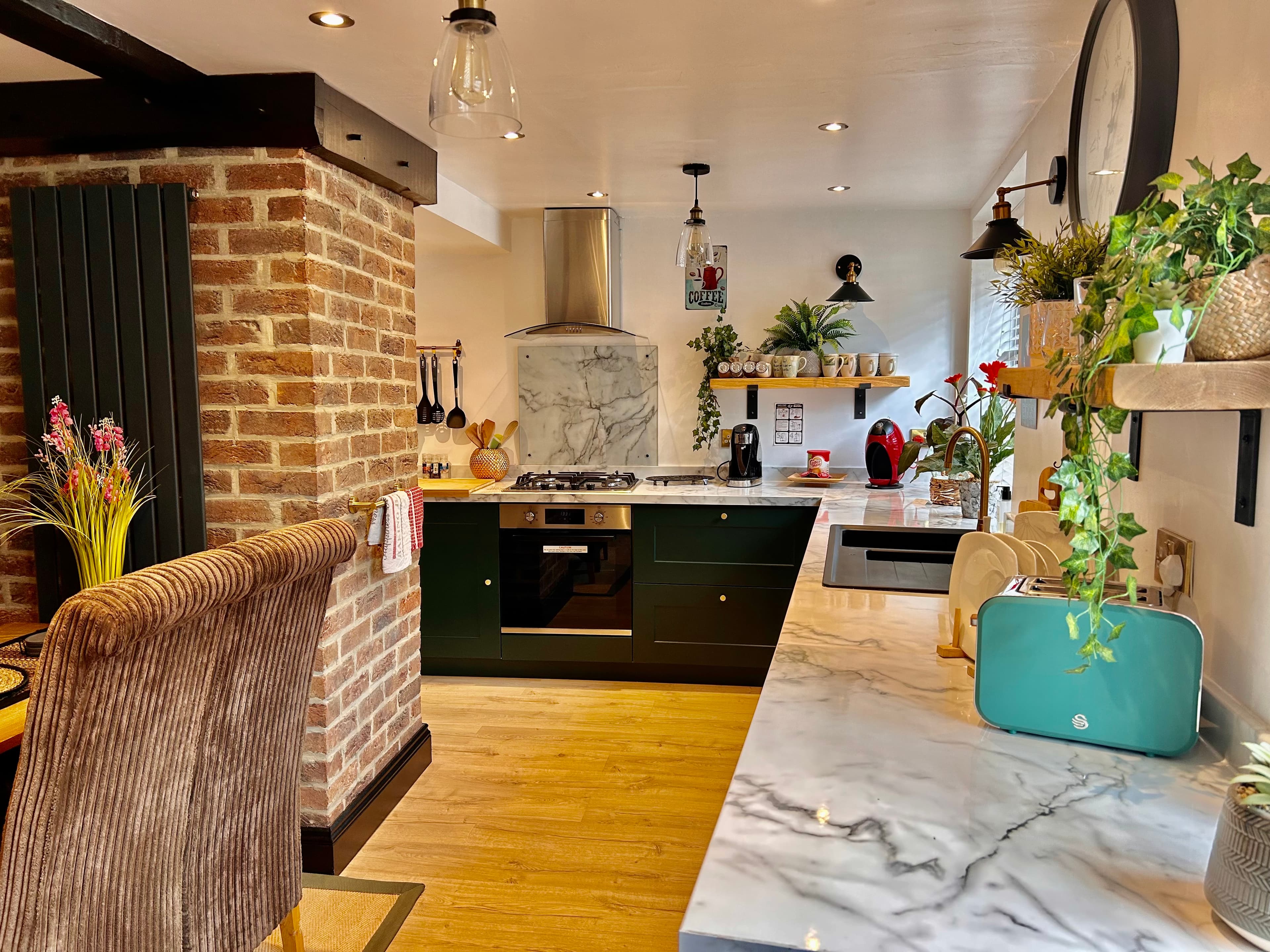 Modern kitchen featuring dark green cabinets, marble countertops, exposed brick, and light wood flooring.