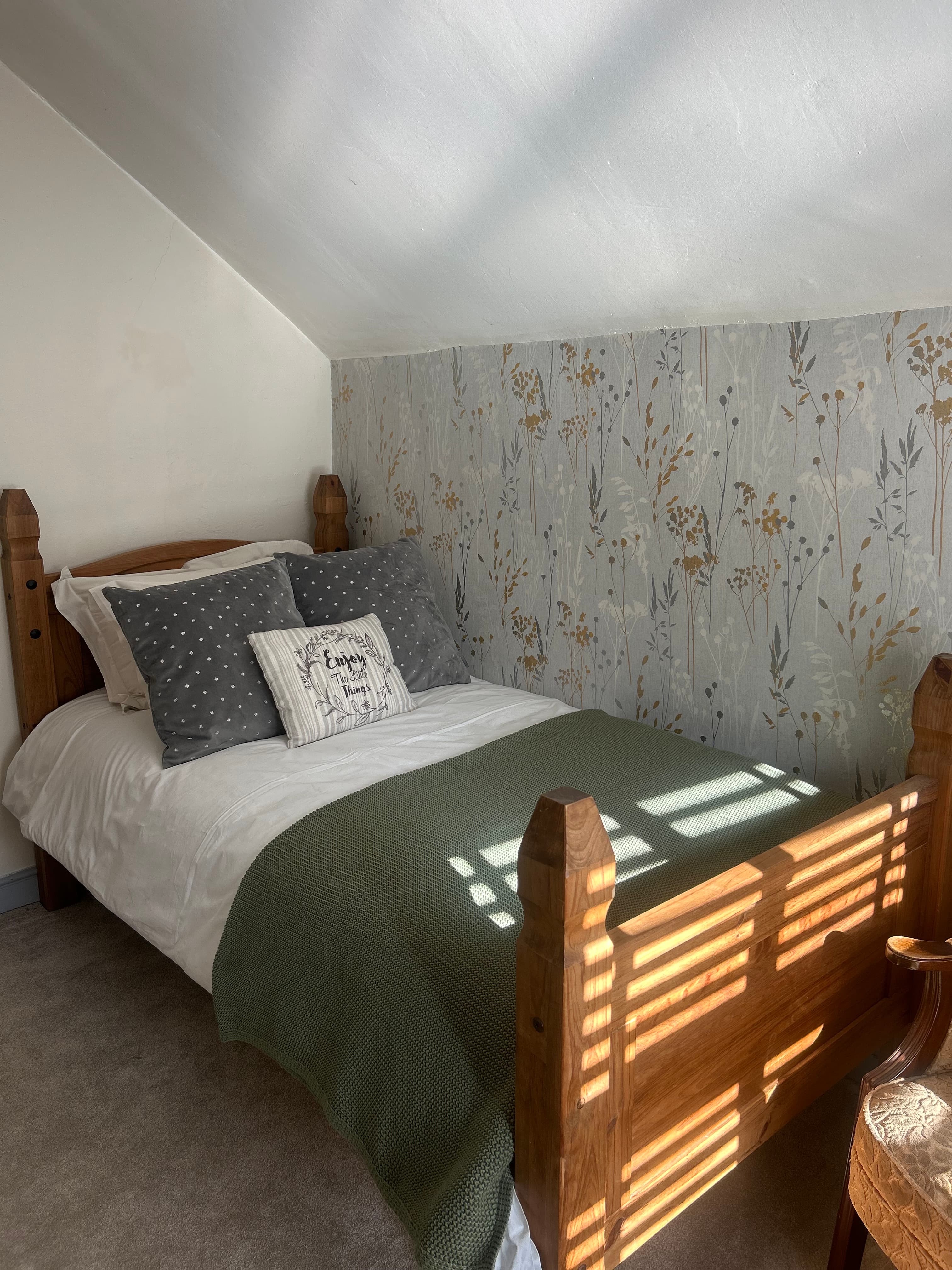 Wooden bed with green throw and floral wallpaper in a sunlit attic bedroom.