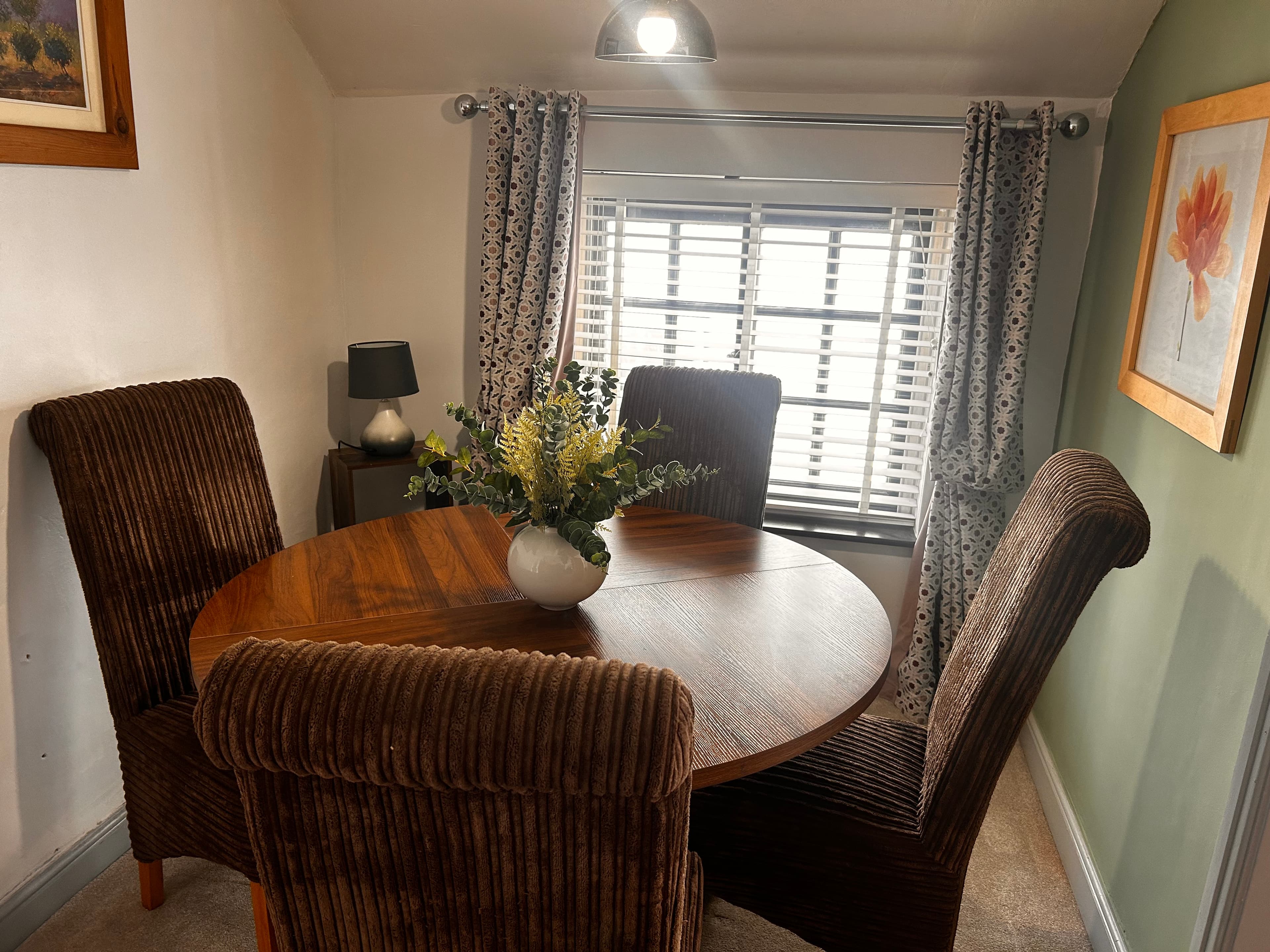 Round wooden table with brown corduroy chairs and a floral centerpiece near a window.