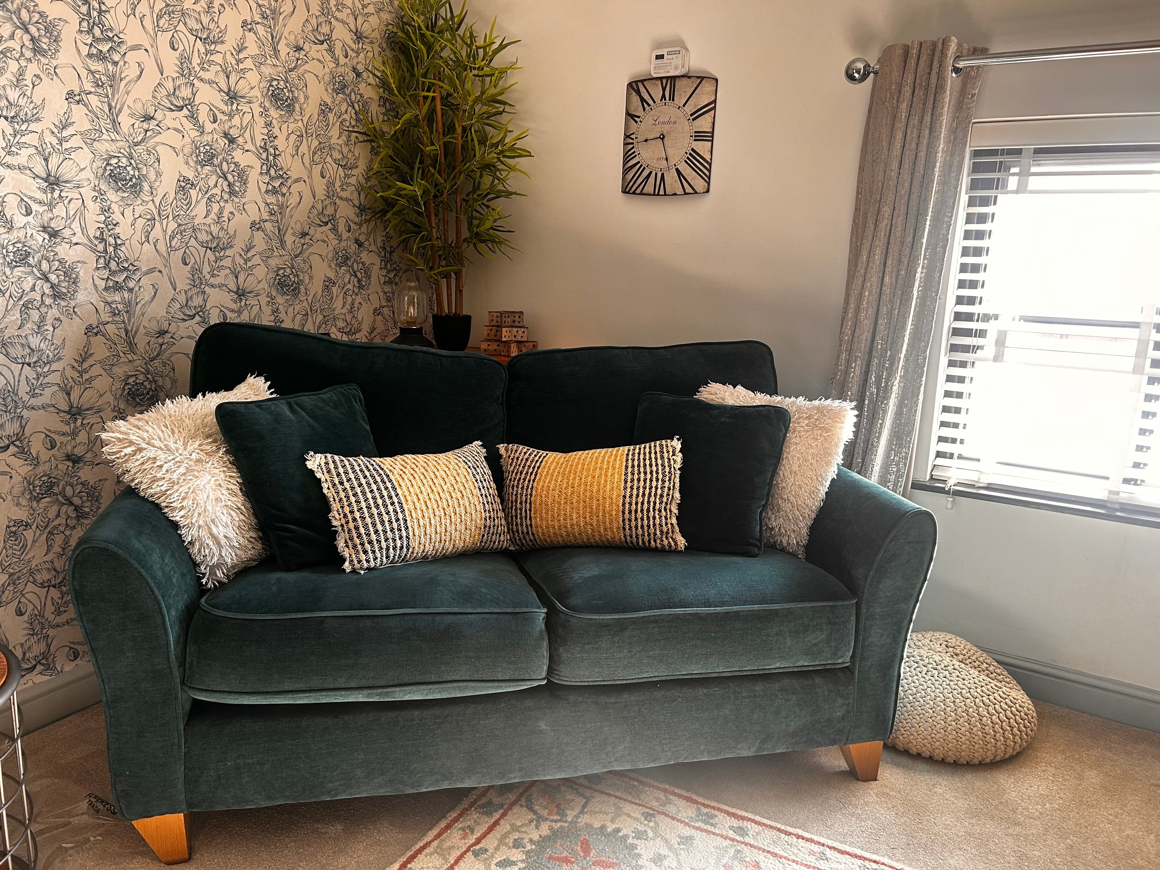 Dark green velvet sofa with decorative pillows, floral wallpaper, a plant, and wall clock.