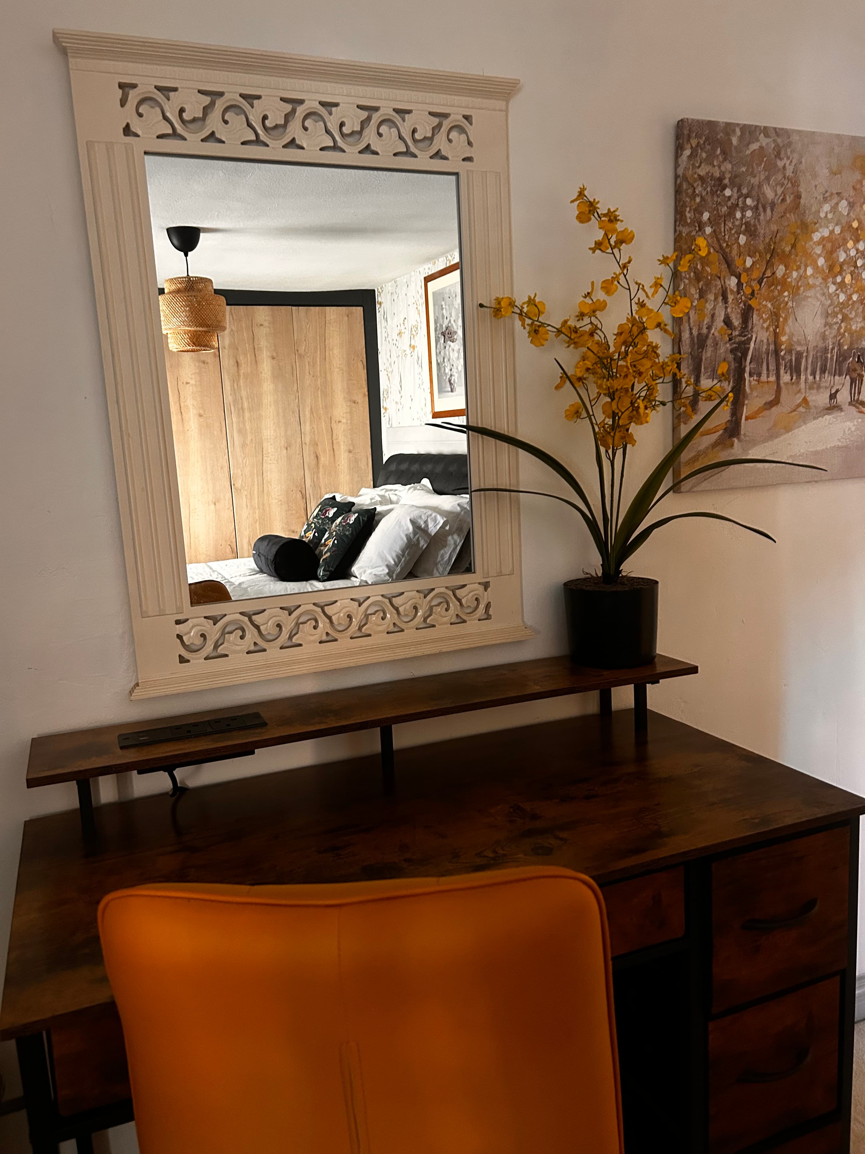 Wooden desk with orange chair, ornate mirror reflecting a bedroom, and yellow potted flowers.